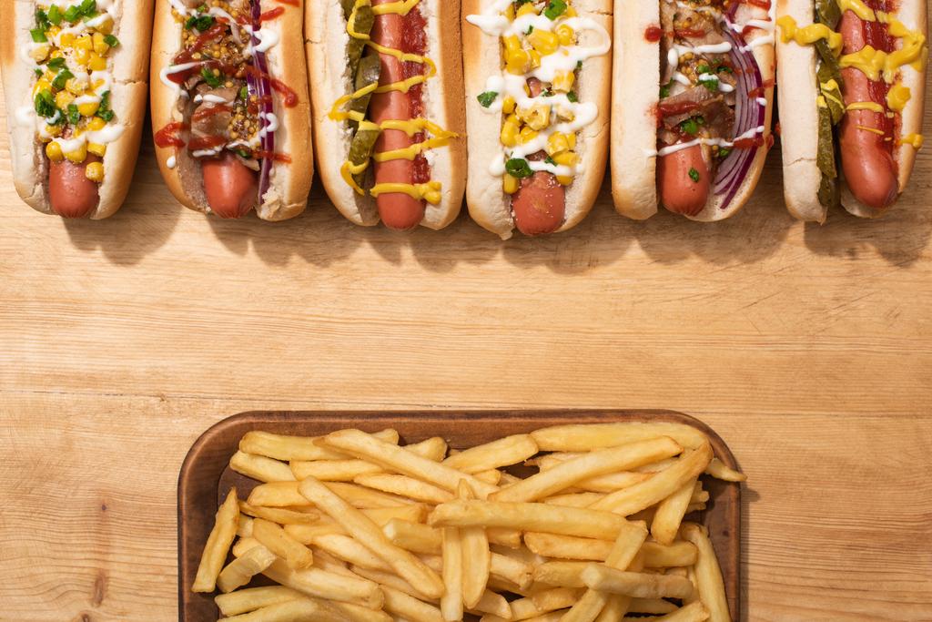 bovenaanzicht van diverse heerlijke hotdogs met groenten en sauzen bij frites op houten tafel - Foto, afbeelding