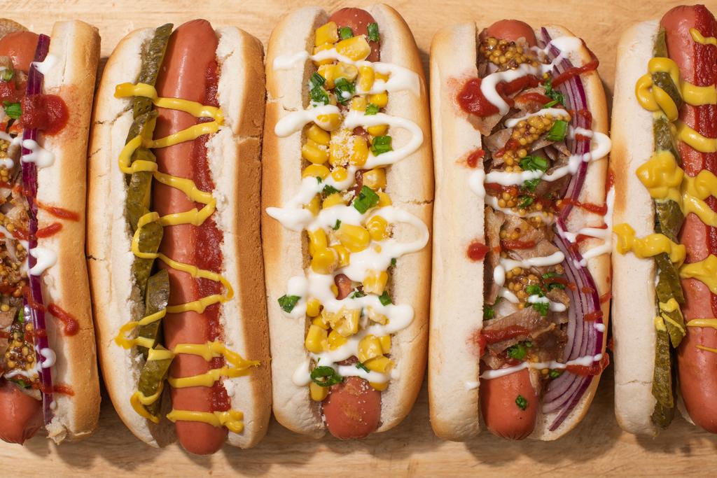 vrchní pohled na různé lahodné hot dogy se zeleninou a omáčkami na dřevěném stole - Fotografie, Obrázek