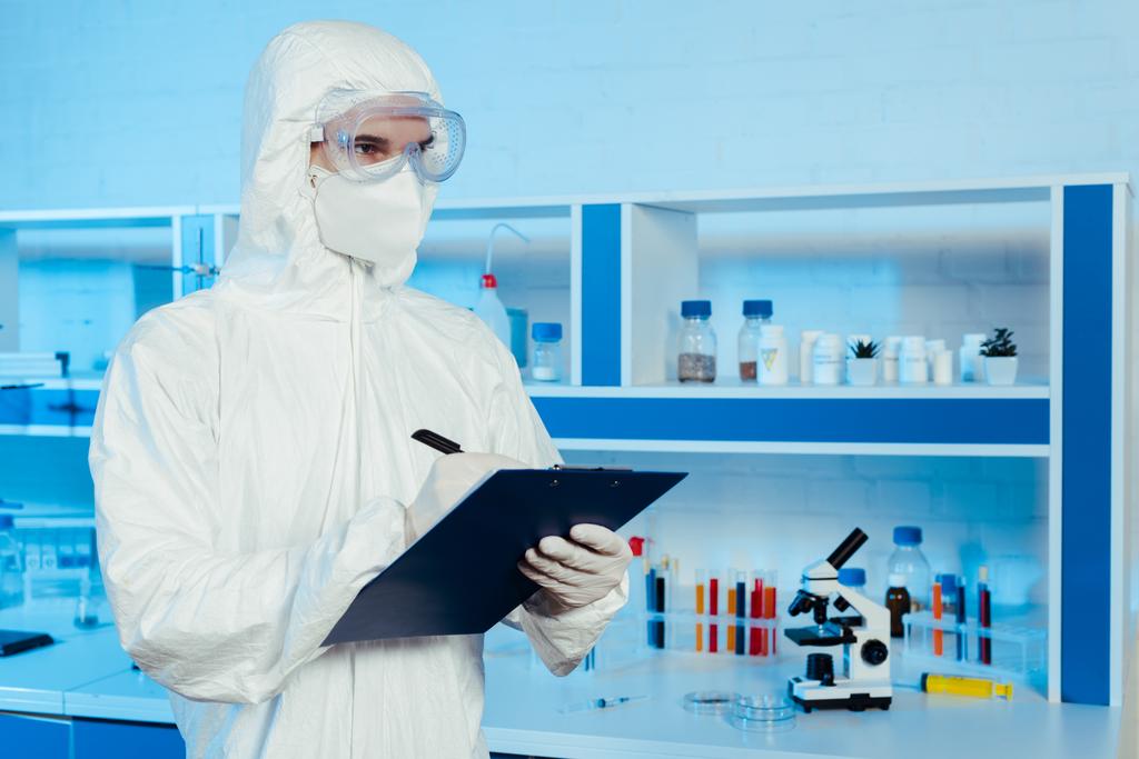 Tehlikeli madde giysisi ve gözlüklü bilim adamı elinde not panosu ve kalemle mikroskobun yanında.  - Fotoğraf, Görsel