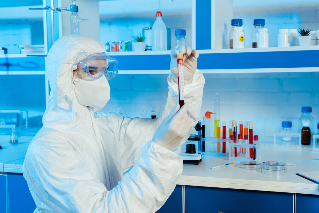 учёный в защитном костюме и латексных перчатках держит пробирку с коронавирусной надписью
  - Фото, изображение