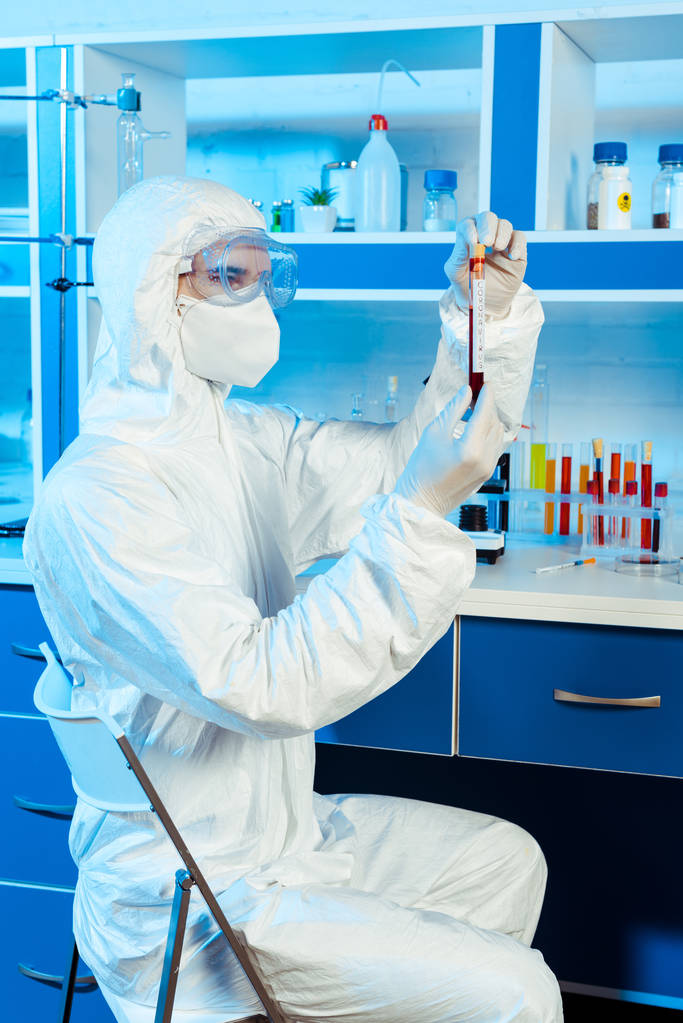 scientist in hazmat suit and latex gloves holding test tube with sample and coronavirus lettering  - Photo, Image