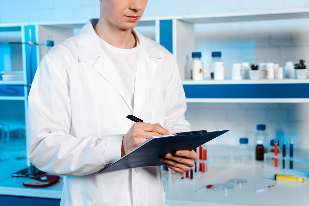 Ausgeschnittene Ansicht eines Wissenschaftlers im weißen Kittel mit Klemmbrett und Stift im Labor  - Foto, Bild