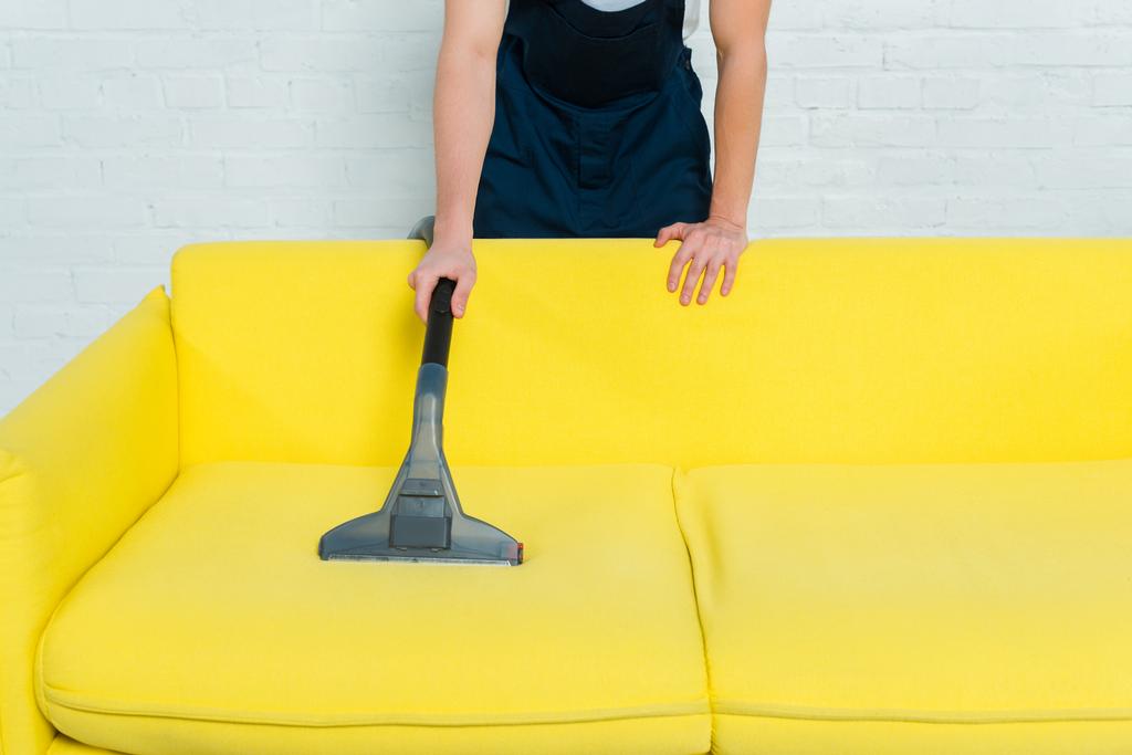 vista ritagliata dell'uomo in uniforme rimozione polvere sul divano con aspirapolvere
  - Foto, immagini