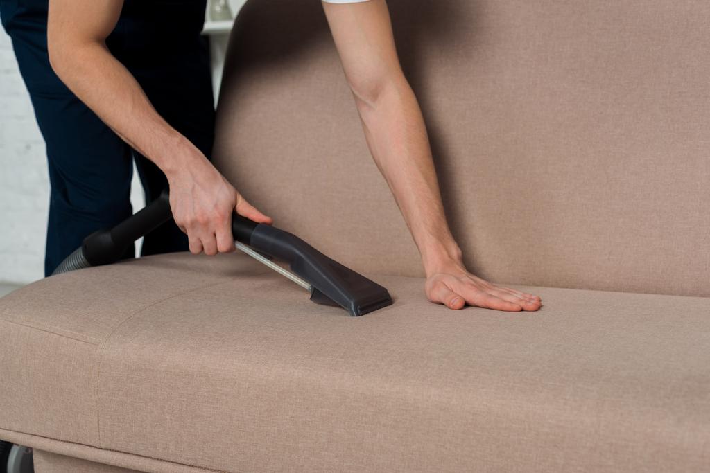 abgeschnittene Ansicht des Mannes, der modernes Sofa mit Staubsauger reinigt  - Foto, Bild