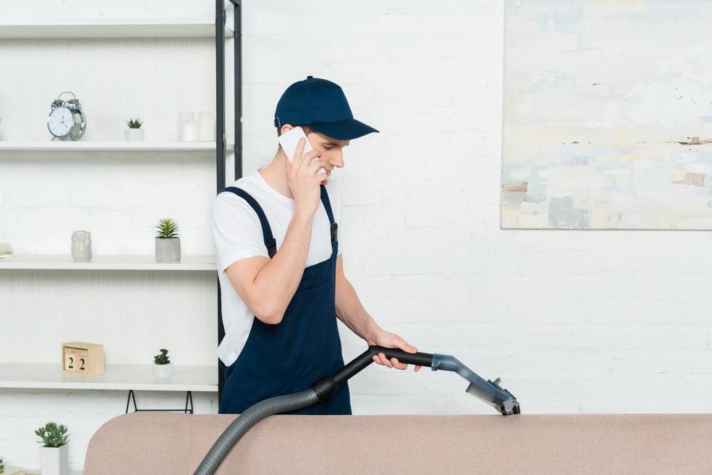 cleaner in cap and overalls cleaning sofa with vacuum cleaner and talking on smartphone  - Photo, Image