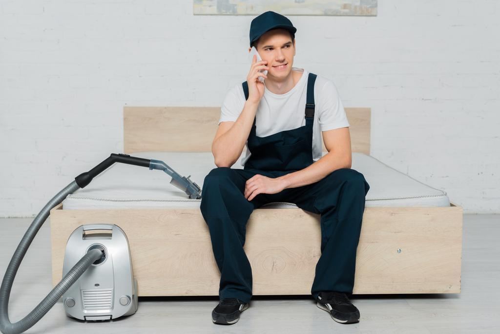 happy cleaner in cap sitting on bed and talking on smartphone near vacuum cleaner  - Photo, Image