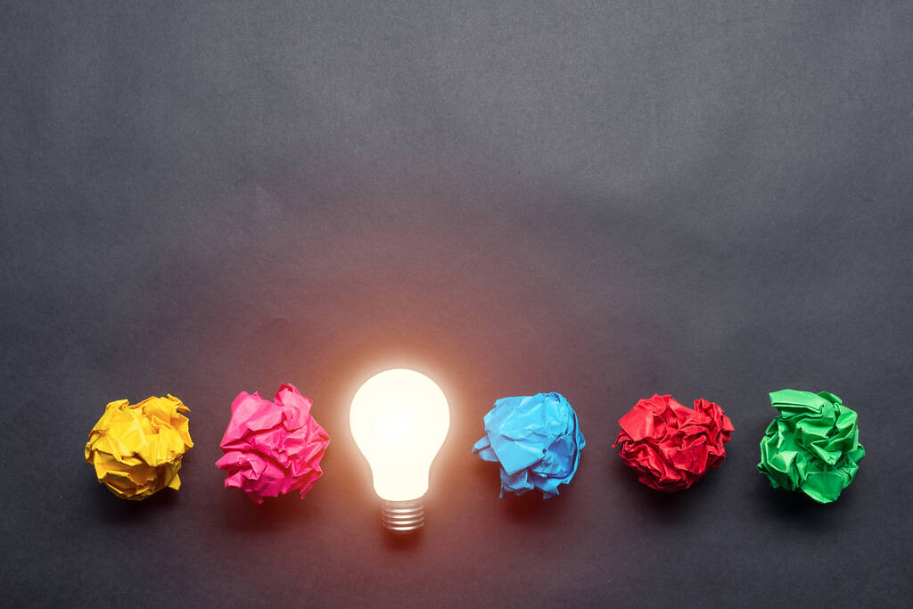 Lightbulb and crumpled colorful paper balls on black background. Successful solution of problem. Idea generation and brainstorming. Genius idea among failing ideas metaphor. Business motivation - Photo, Image