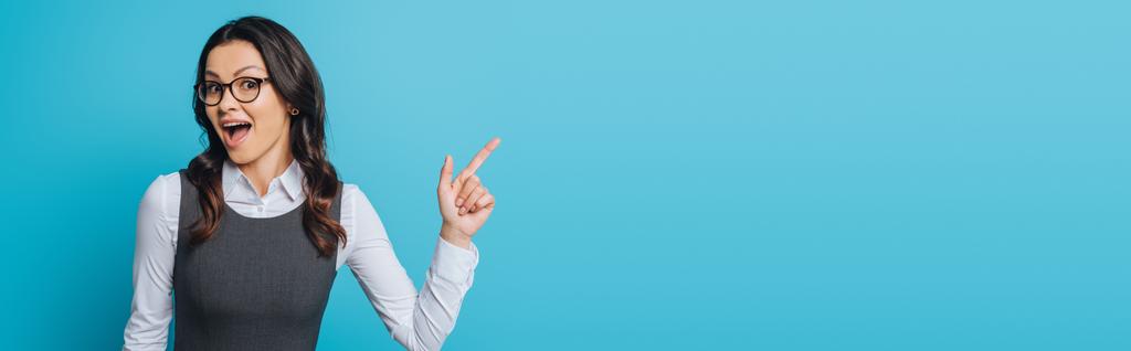 tiro panorâmico de empresária maravilhada em óculos apontando com dedo isolado em azul
 - Foto, Imagem