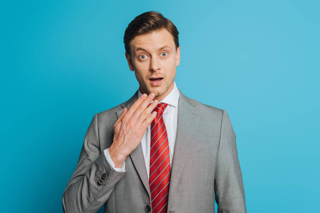 shocked businessman touching chin while looking at camera with wide open eyes on blue background - Photo, Image