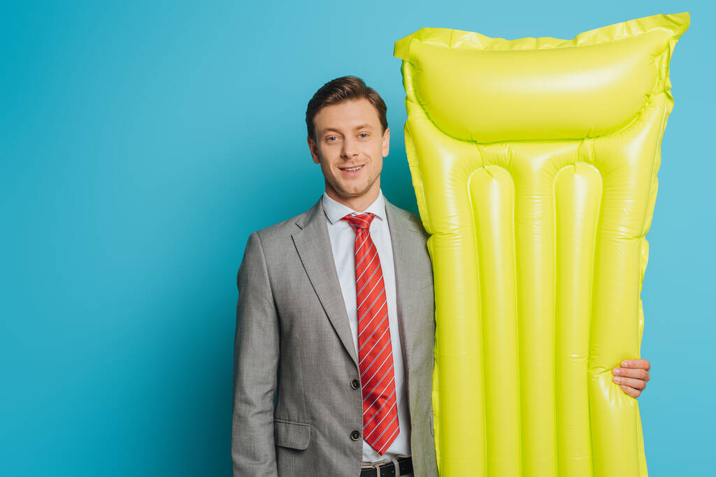 feliz hombre de negocios que sostiene el colchón inflable mientras que mira la cámara en fondo azul
 - Foto, imagen
