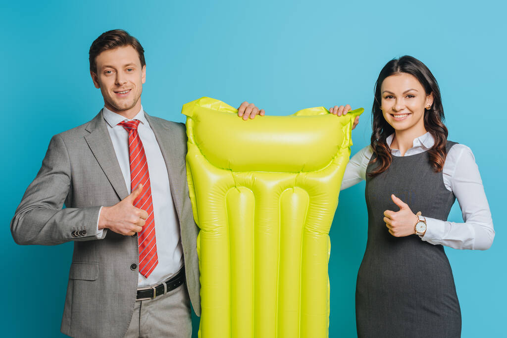 δύο ευτυχισμένοι επιχειρηματίες κρατώντας κίτρινο φουσκωτό στρώμα και δείχνοντας αντίχειρες επάνω στο μπλε φόντο - Φωτογραφία, εικόνα