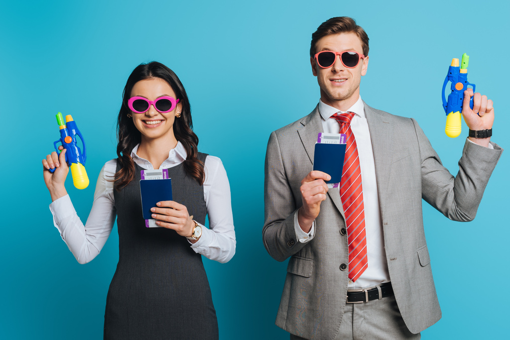 dois empresários felizes em óculos de sol segurando armas de água, passaportes e passagens aéreas em fundo azul
 - Foto, Imagem