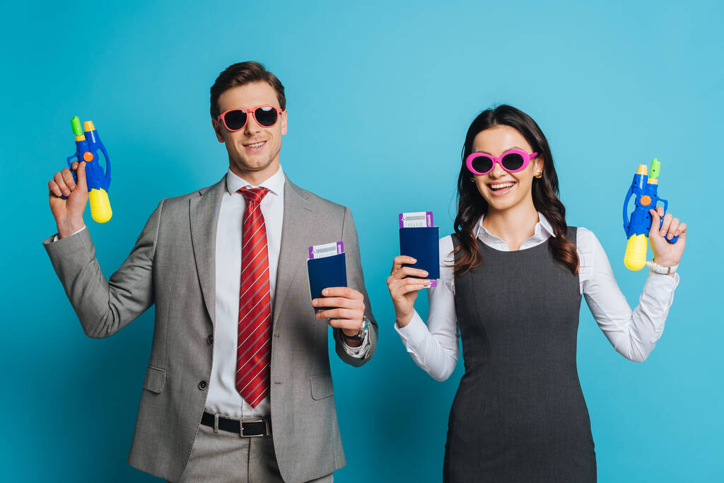 allegro uomo d'affari e donna d'affari in occhiali da sole in possesso di pistole ad acqua, passaporti e biglietti aerei su sfondo blu
 - Foto, immagini