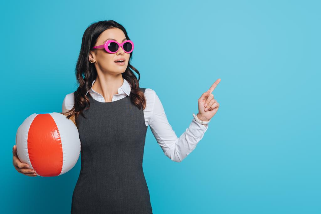 удивлённая деловая женщина в солнечных очках, держащая надувной мяч, отводя взгляд и указывая пальцем на синий фон
 - Фото, изображение