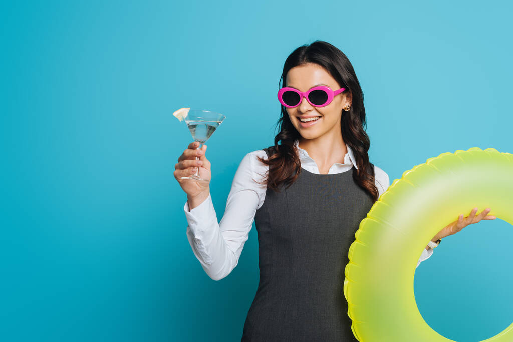 cheerful businesswoman in sunglasses holding glass of cocktail and swim ring on blue background - Photo, Image