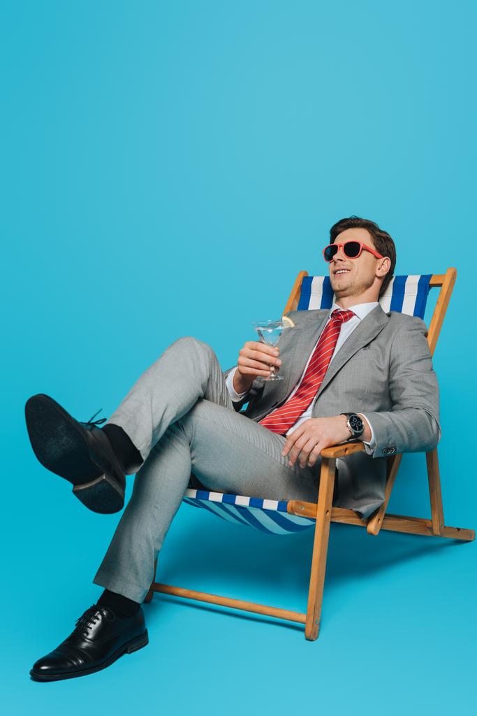 счастливый бизнесмен в солнечных очках сидит в шезлонге со стаканом коктейля на синем фоне
 - Фото, изображение