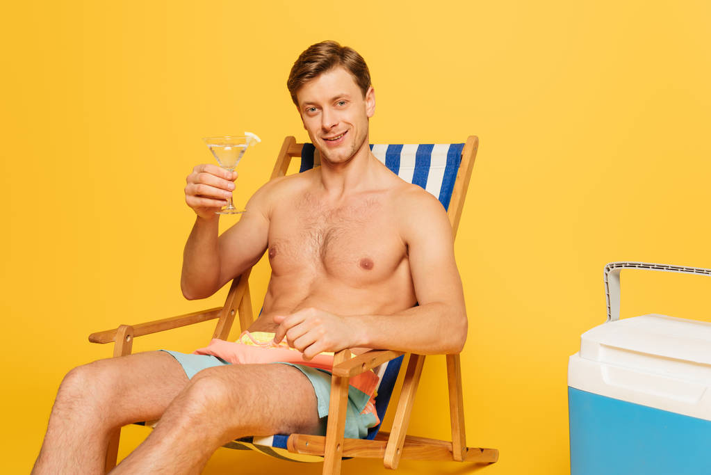 szczęśliwy człowiek bez koszuli siedzi na leżaku obok przenośnej lodówki i trzyma szklankę koktajlu na żółtym tle - Zdjęcie, obraz