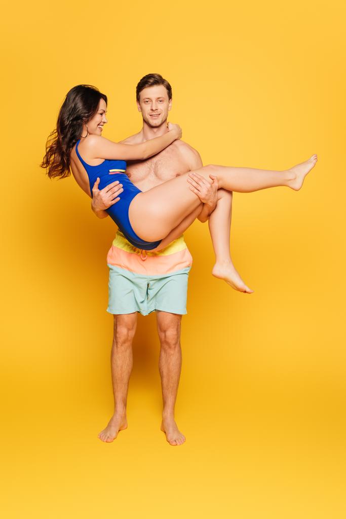 handsome shirtless man holding happy girlfriend in swimsuit on yellow background - Photo, Image