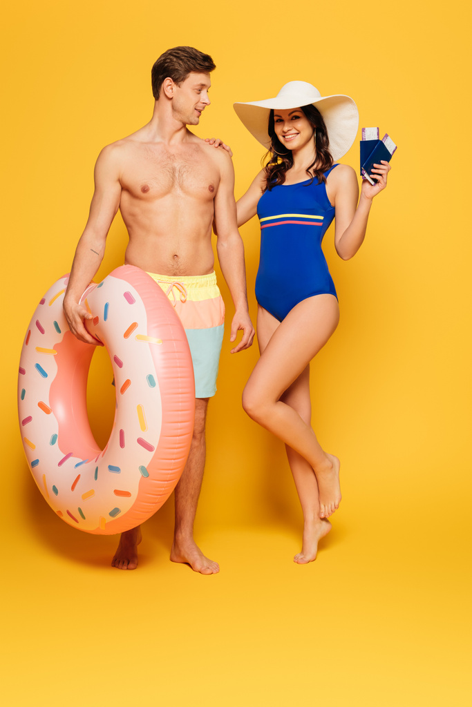 glückliche Frau im Badeanzug, die Pässe und Flugtickets in der Hand hält, während sie die Schulter eines hemdlosen Mannes mit aufblasbarem Ring auf gelbem Hintergrund berührt - Foto, Bild