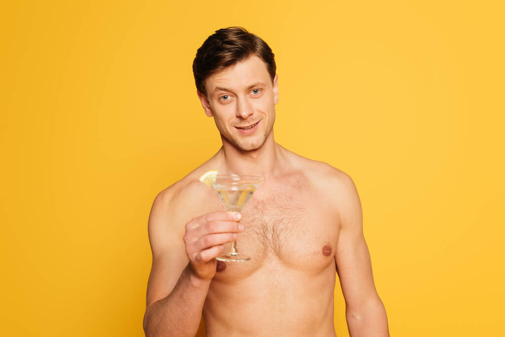 красивий без сорочки чоловік дивиться на камеру, тримаючи склянку коктейлю ізольовано на жовтому
 - Фото, зображення