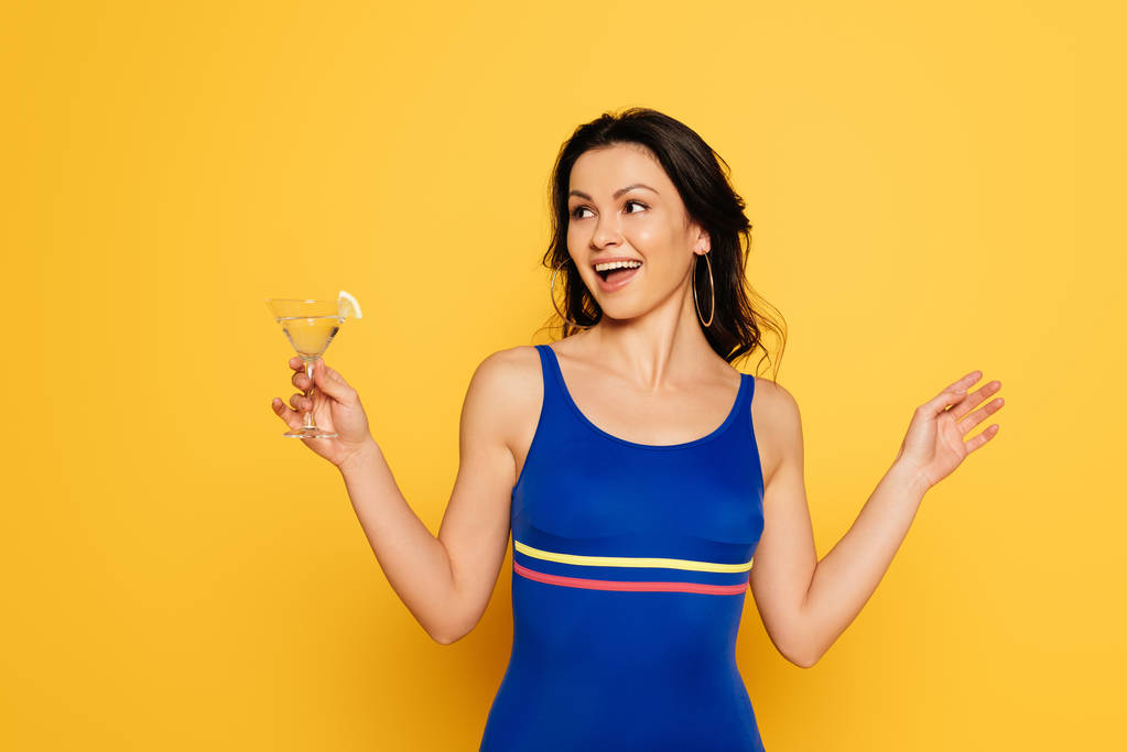 весела жінка в синьому купальнику тримає склянку коктейлю і дивиться ізольовано на жовтий
 - Фото, зображення