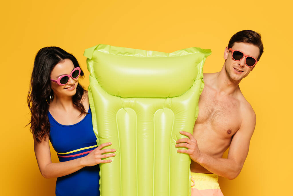 fröhliches Paar mit Sonnenbrille und aufblasbarer Matratze auf gelbem Hintergrund - Foto, Bild
