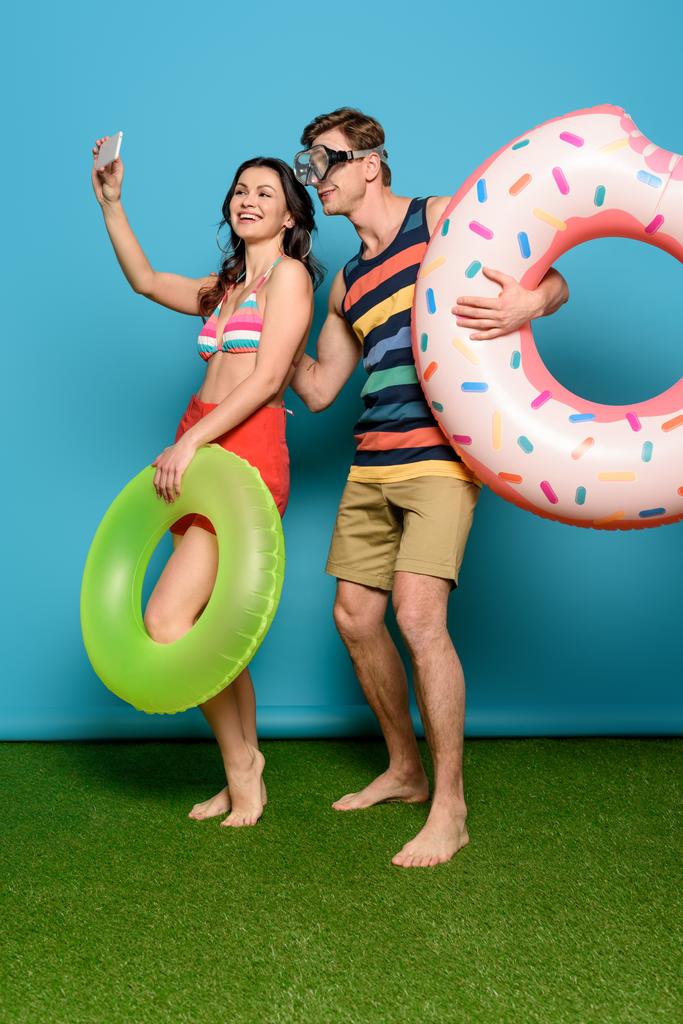 alegre pareja con anillos inflables tomando selfie en teléfono inteligente sobre fondo azul
 - Foto, imagen