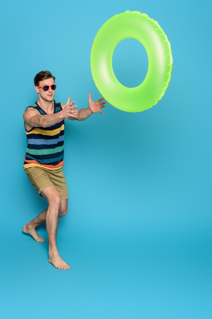 Çizgili atlet ve şortlu genç adam mavi arka planda şişme yüzük atıyor. - Fotoğraf, Görsel