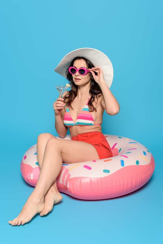 κομψή γυναίκα με γυαλιά ηλίου και καπέλο ήλιο κάθεται στο δαχτυλίδι κολύμπι, ενώ κρατώντας ένα ποτήρι κοκτέιλ σε μπλε φόντο - Φωτογραφία, εικόνα