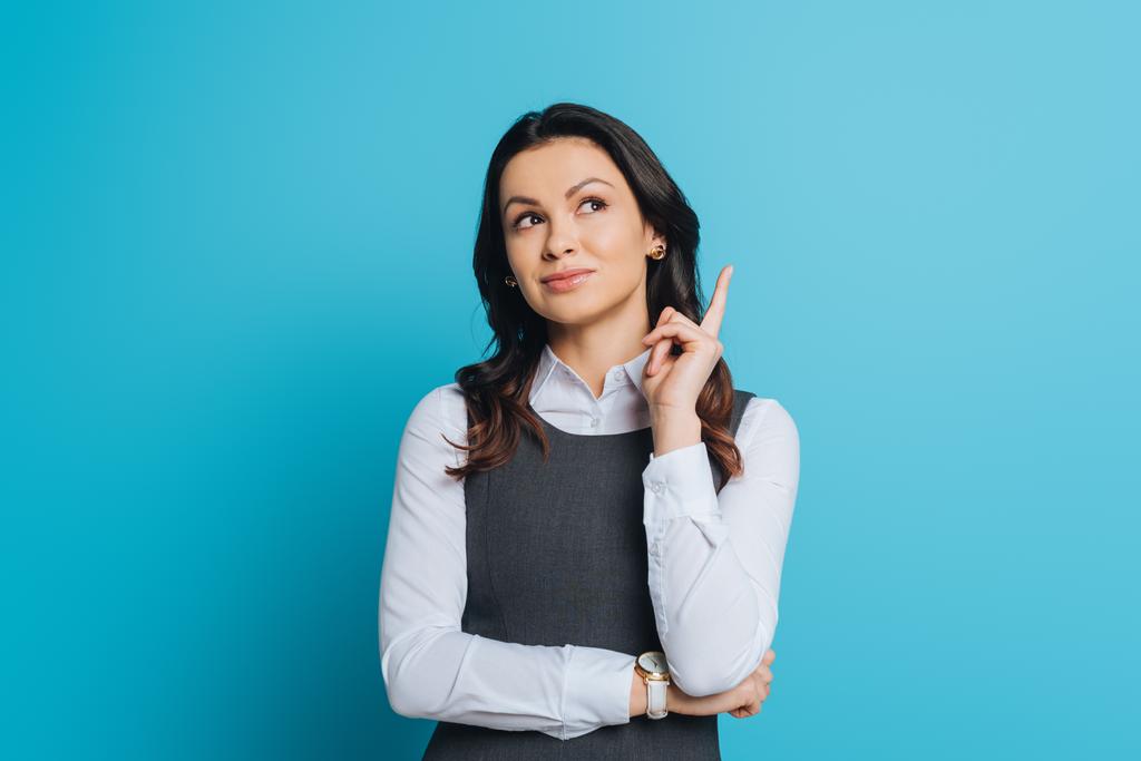 dreamy businesswoman looking up while showing idea gesture on blue background - Photo, Image