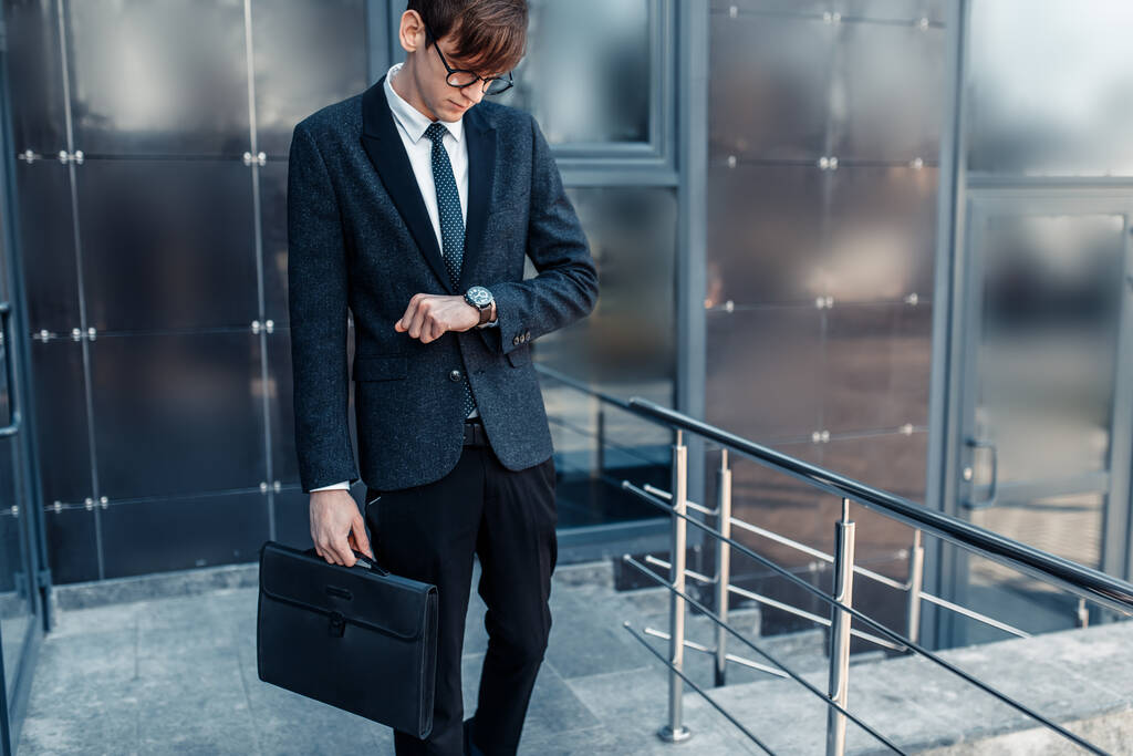 Стильный красивый молодой бизнесмен в деловом костюме, опаздывающий на работу смотрит на часы на руке, стоя на фоне стеклянного здания на улице
 - Фото, изображение