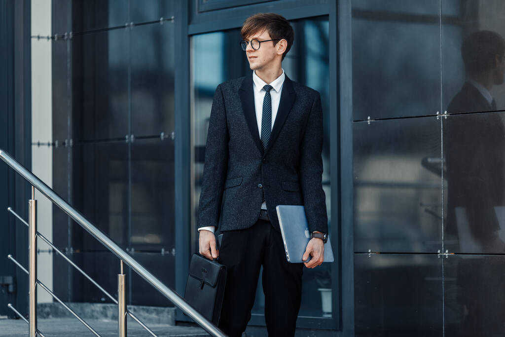 Привлекательный серьезный молодой бизнесмен в деловом костюме, держащий ноутбук на улице на фоне стеклянного офиса
 - Фото, изображение