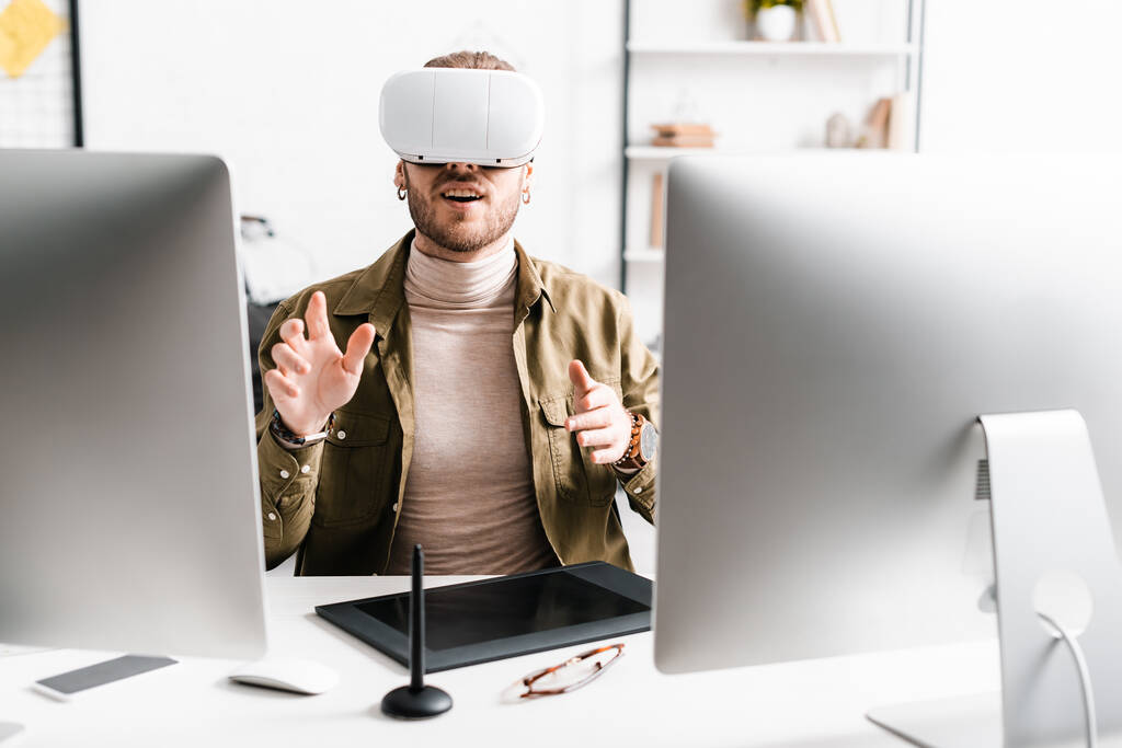 Podekscytowany 3D artysta w wirtualnej rzeczywistości zestaw słuchawkowy gestykulacji w pobliżu komputerów i tabletów graficznych na stole - Zdjęcie, obraz