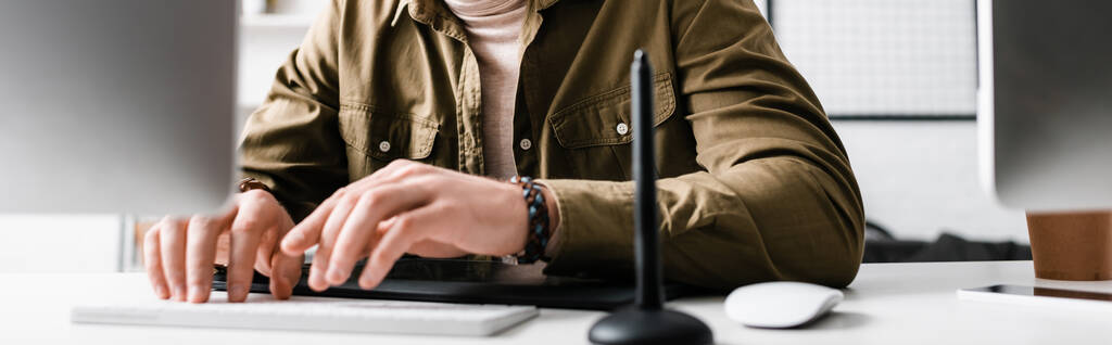 Vista recortada del diseñador digital escribiendo en el teclado de la computadora cerca de la tableta gráfica en la mesa, plano panorámico
 - Foto, imagen