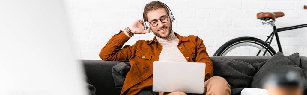 Выборочный фокус улыбающегося 3d артиста, слушающего музыку и использующего ноутбук на диване в офисе, панорамный снимок
 - Фото, изображение