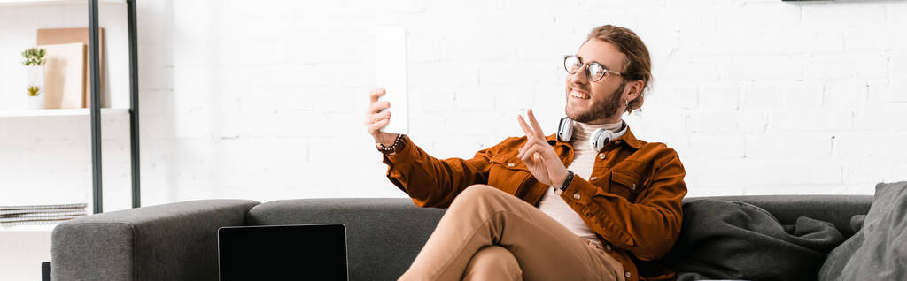 Foto panorámica de sonriente artista 3d mostrando signo de paz mientras que tiene videollamada en tableta digital cerca de portátil en sofá
  - Foto, imagen