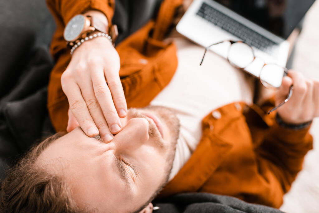 Widok na zmęczonego projektanta 3D dotykając oka i trzymając okulary z laptopem na kanapie   - Zdjęcie, obraz
