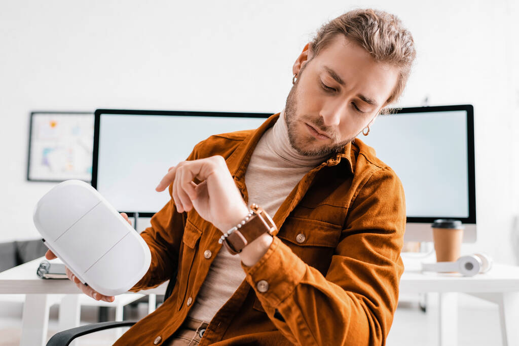 Przystojny 3D artysta patrząc na zegarek i trzymając zestaw słuchawkowy VR w biurze  - Zdjęcie, obraz