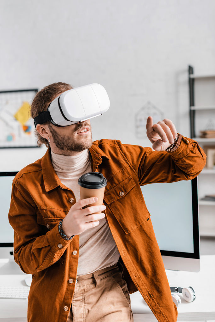 Designer digital apontando com o dedo enquanto usa headset realidade virtual e segurando copo de papel no escritório
 - Foto, Imagem