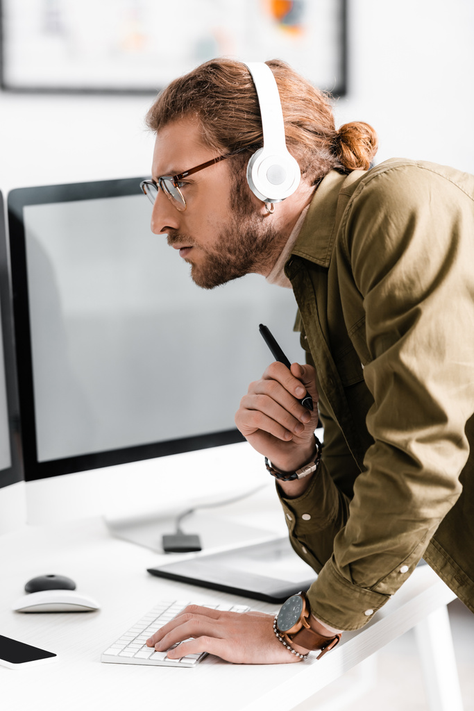 Widok z boku przystojny projektant cyfrowy w słuchawkach za pomocą komputera i trzymając rysik tabletu graficznego przy stole w biurze  - Zdjęcie, obraz