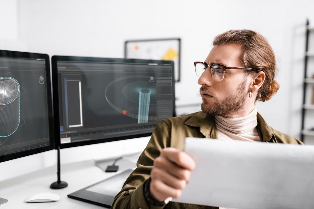 Selective focus of digital designer holding blueprint and looking project of 3d design on computer monitors on table  - Photo, Image