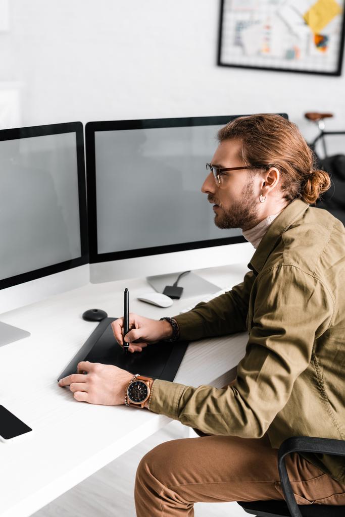Widok boczny przystojnego projektanta cyfrowego pracującego z tabletem graficznym i komputerami z pustym ekranem na stole  - Zdjęcie, obraz