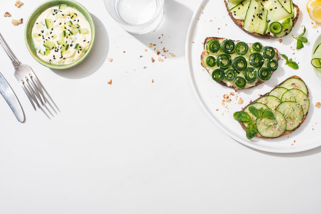 top view of fresh cucumber toasts on plate near lemon, water, cutlery and yogurt on white background - Photo, Image