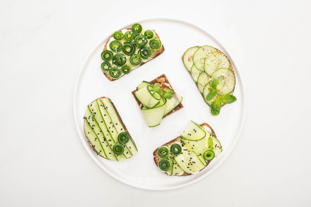 Yuvarlak tabakta tohum, nane ve fesleğen yapraklı salatalık tostlarının beyaz arka planda üst görüntüsü - Fotoğraf, Görsel