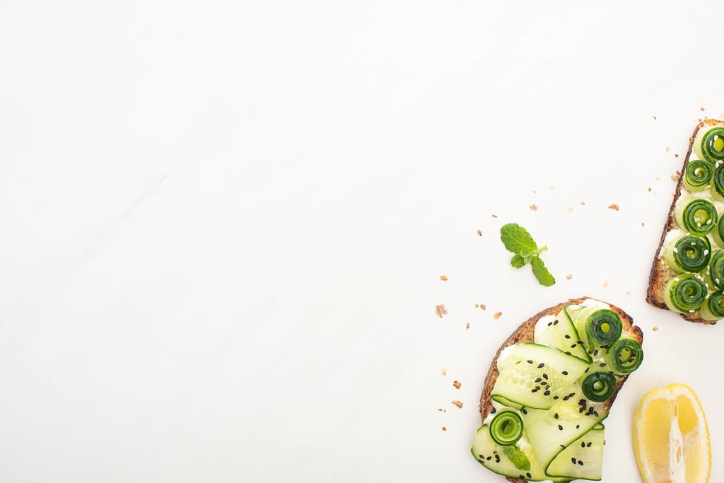 bovenaanzicht van verse komkommer toast met zaden, munt en basilicum bladeren en citroen op witte achtergrond - Foto, afbeelding