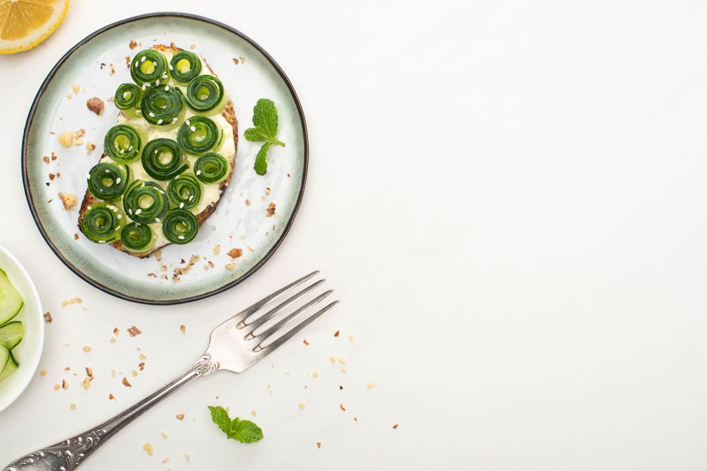 bovenaanzicht van verse komkommer toast met sesam en munt bladeren op plaat in de buurt van vork en citroen op witte achtergrond - Foto, afbeelding