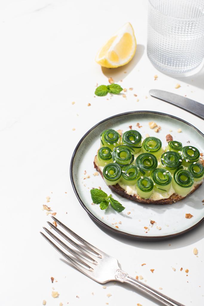 verse komkommer toast met zaden geserveerd met bestek, citroen en water op witte achtergrond - Foto, afbeelding