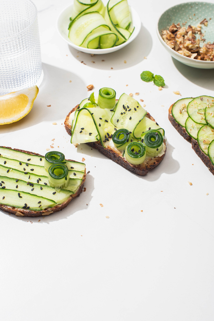 verse komkommer toast met zaden, munt bladeren en citroen in de buurt van glas water op witte achtergrond - Foto, afbeelding