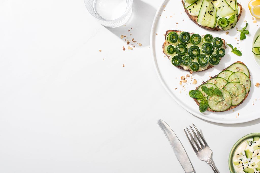 top view of fresh cucumber toasts with seeds, mint and basil leaves on plate near cutlery, water and yogurt on white background - Photo, Image