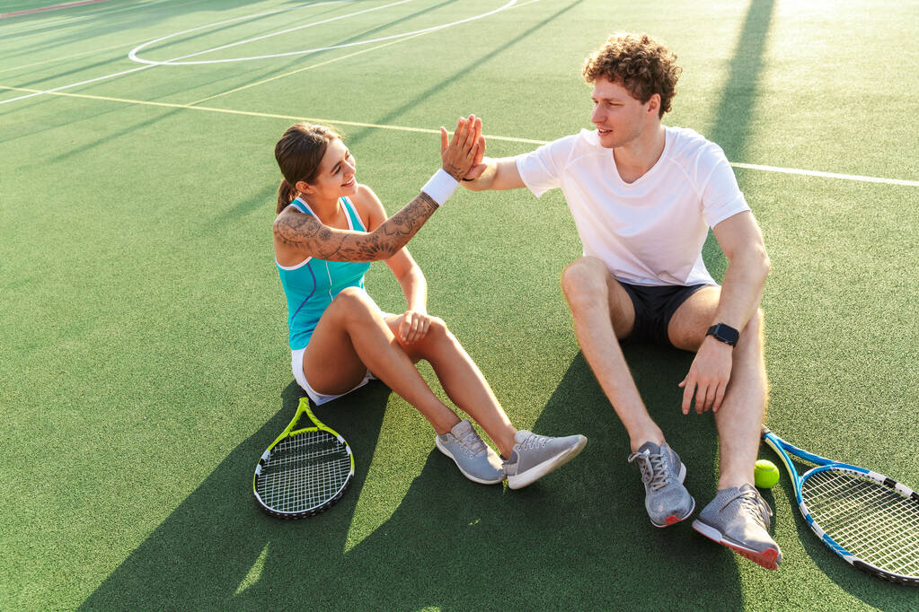 Bild des schönen Mannes und der schönen Frau in Sportbekleidung, die mit Schlägern auf dem Platz sitzen, während sie draußen Tennis spielen - Foto, Bild